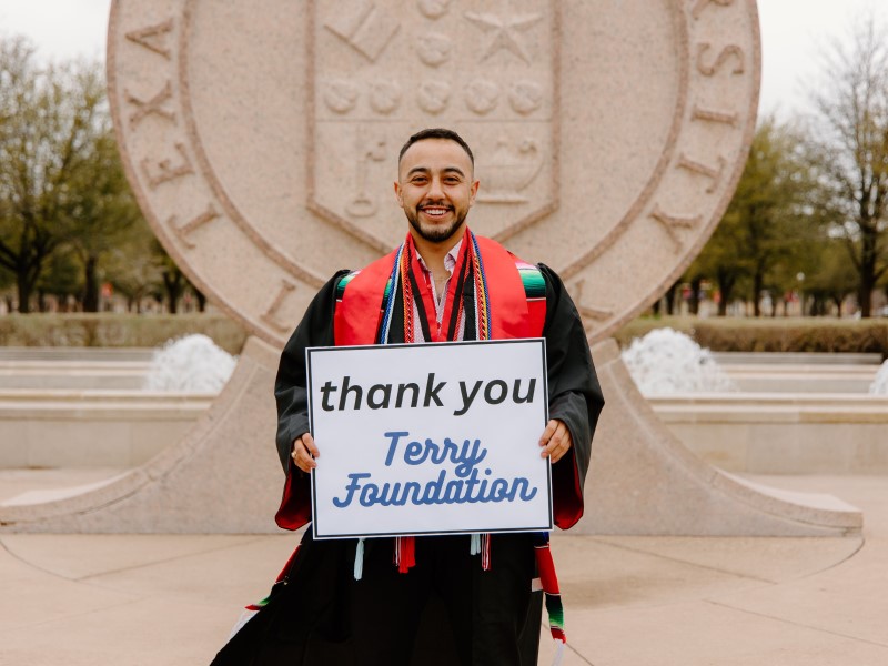 A young man in graduation regalia holding a sign that says "Thank You Terry Foundation"